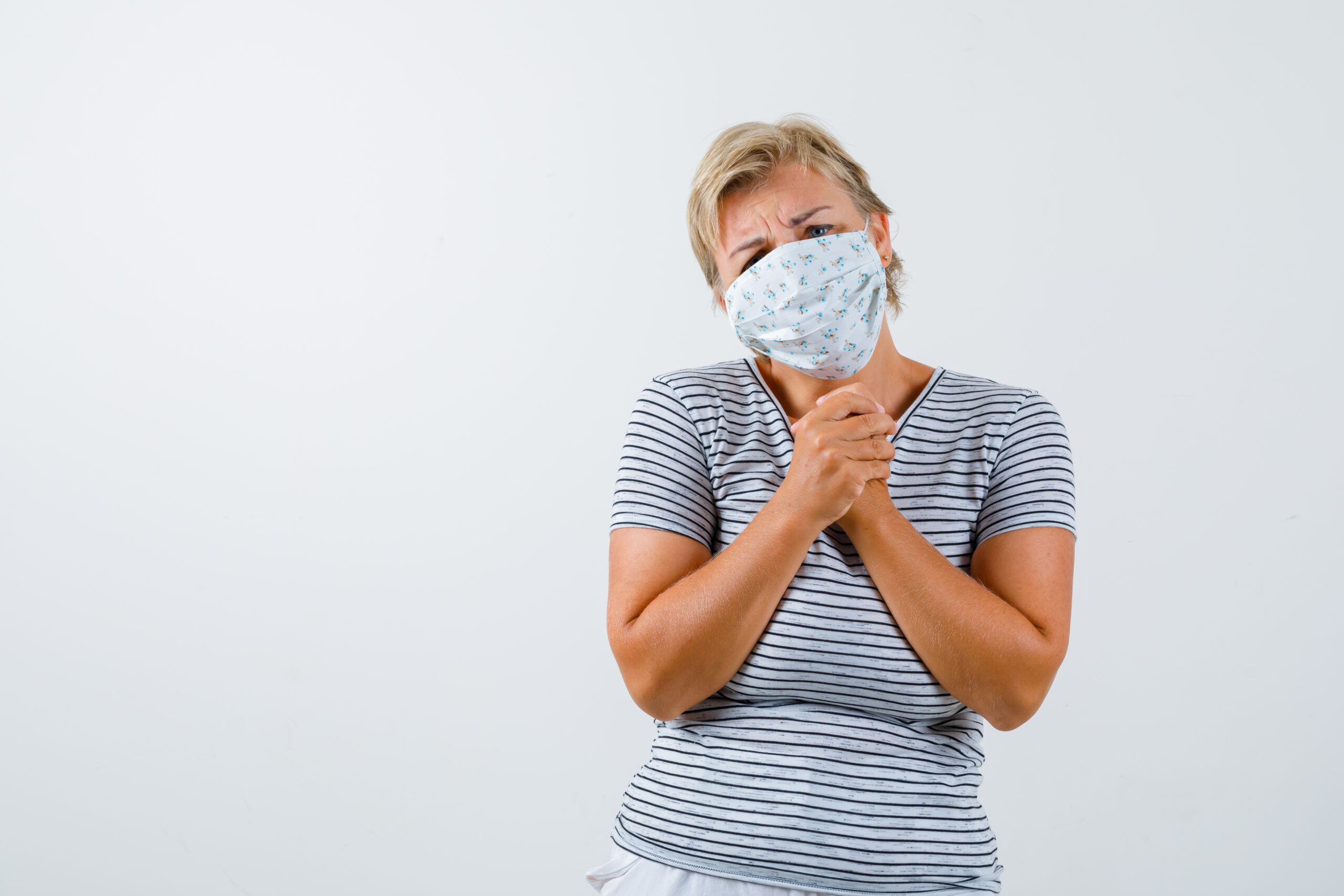 Portrait of blonde mature woman clasping hands in praying gesture in striped t-shirt, mask and looking hopeful front view